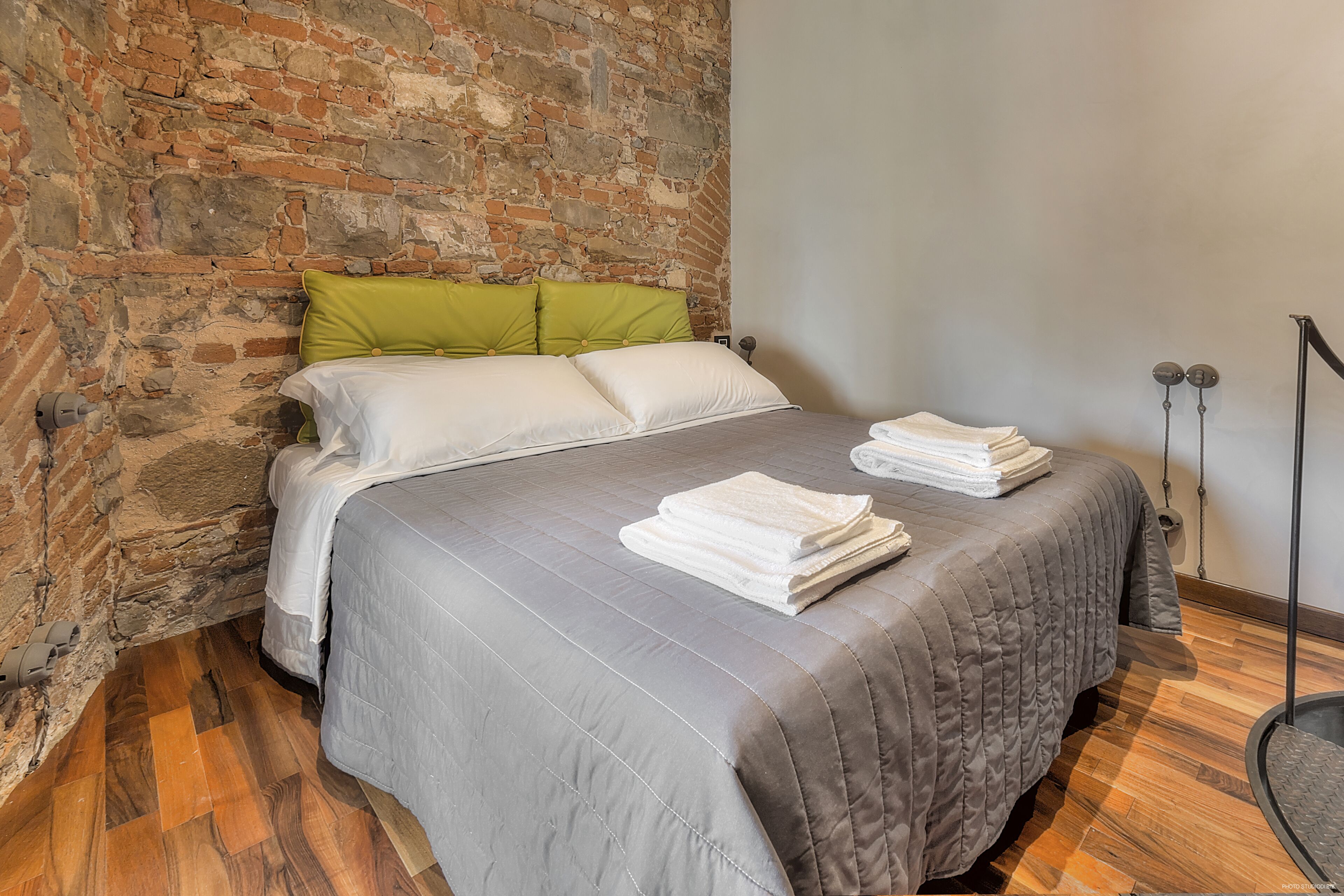 A room with a brick wall, a bed with grey bedding, and a wooden floor.