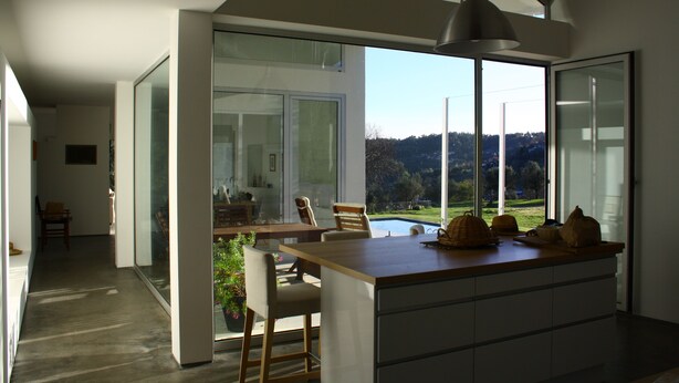 A modern kitchen with a large island, a dining area with chairs, and a view of a swimming pool and hills. - 4 bedroom house with private pool (Celorico de Basto)