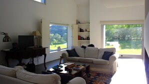 A modern living room with a sofa, coffee table, and a television on a stand. There is a bookshelf with books and decorative items, and a large window offering a view of greenery outside. - 4 bedroom house with private pool (Celorico de Basto)
