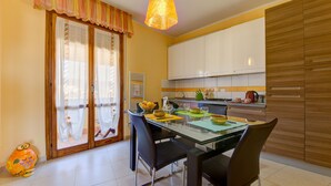 Un coin repas avec une table en verre, des chaises, un luminaire suspendu et une cuisine en arrière-plan.