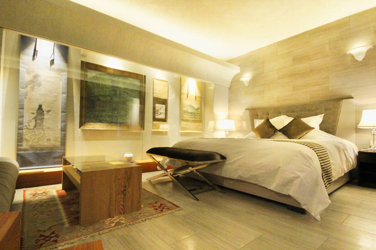 Una habitación de hotel moderna con una cama grande, una mesita de noche, una silla y un ventanal amplio.