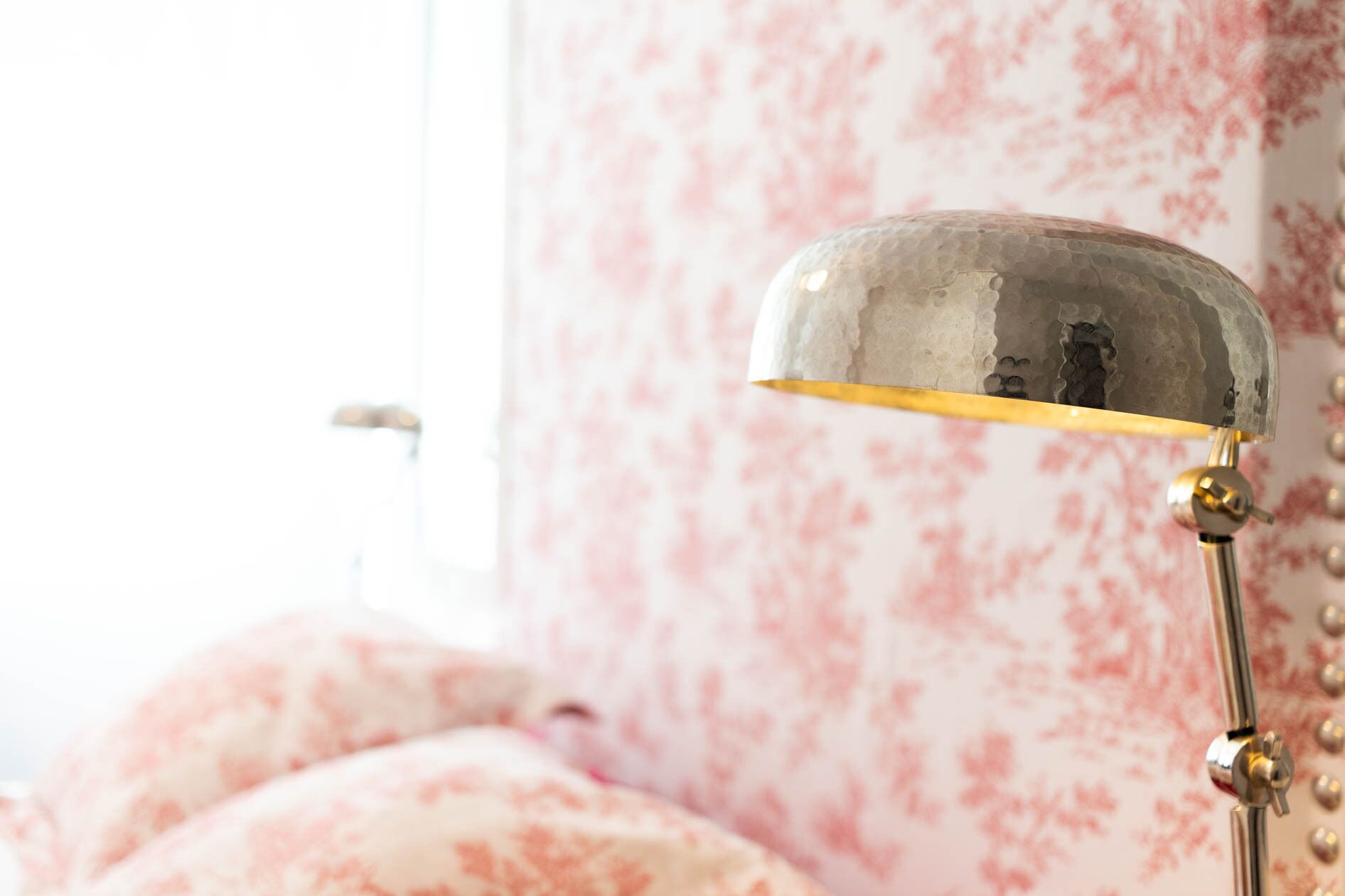 Una lámpara con acabado metálico y base dorada, ubicada frente a una cama con estampado floral.