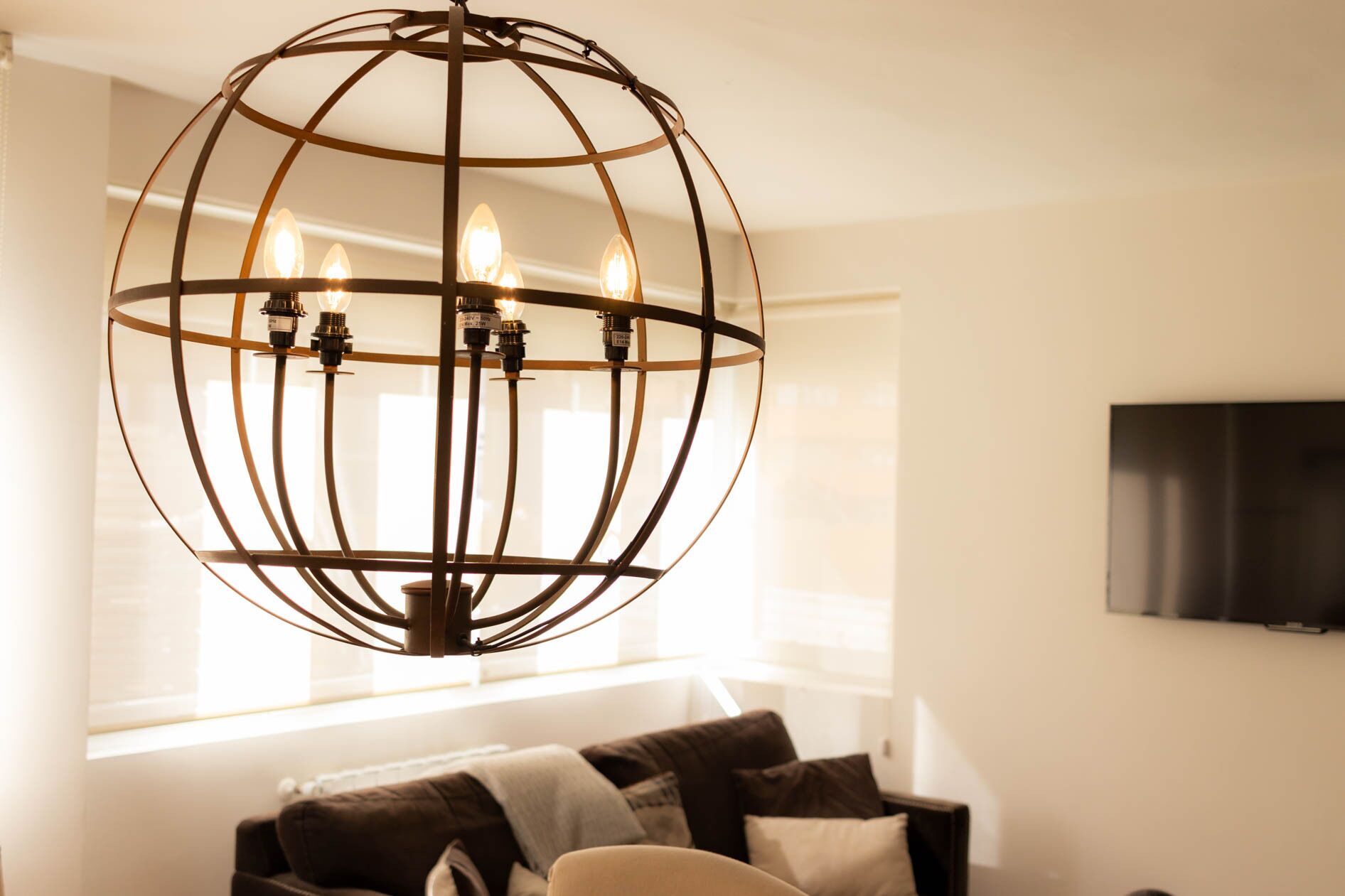 Ein kugelförmiger Kronleuchter mit mehreren Lampen, ein Sofa mit Kissen und ein Fernseher an der Wand.