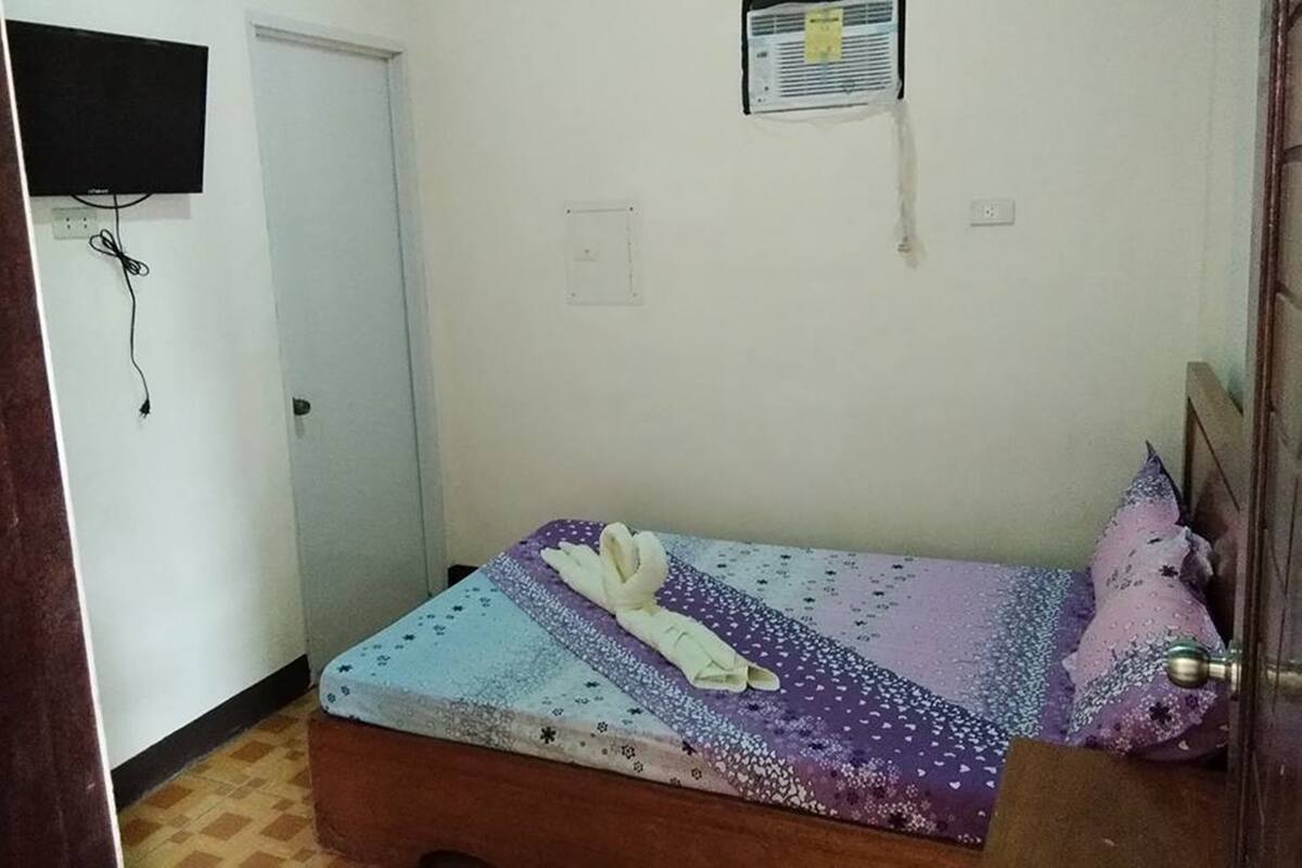 A small, single-bed room with a wooden headboard, a bedspread with a star pattern, a white towel, and a purple pillow. There is a wall-mounted air conditioning unit and a television on the wall.
