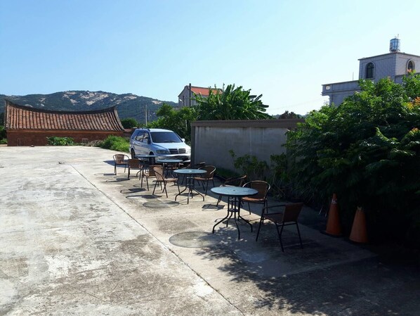 A parking area with a white van, outdoor tables and chairs, and a building with a tiled roof.