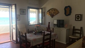Ein Essbereich mit einem für zwei Personen gedecktem Tisch, einem Fernseher auf einer Kommode und Blick auf den Strand durch offene Fenster.