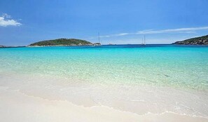 Une plage à l’eau turquoise et limpide, bordée d’un rivage sablonneux, avec des collines verdoyantes à l’horizon.