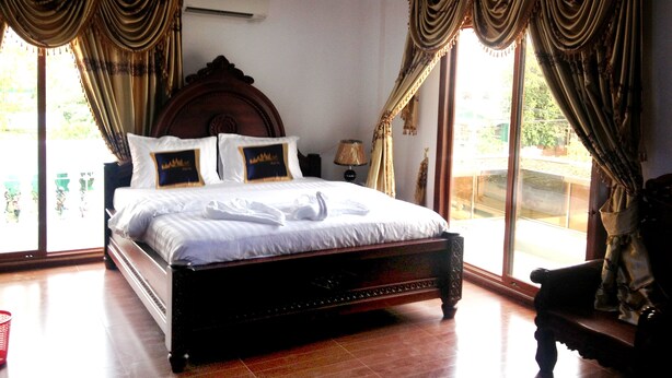 Una cama grande con cabecera de madera, dos almohadas con diseño de cresta y una colcha bien tendida. Se ve una ventana con cortinas, una mesita de noche con lámpara, y una silla en la esquina.