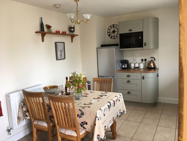A dining area with a table set for four, a chandelier, a clock, and a kitchen with a refrigerator and microwave.