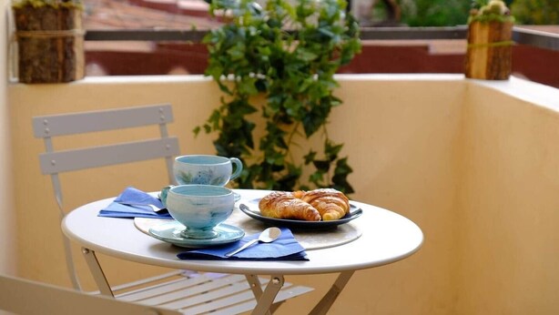 Ein Tisch mit einer Croissantplatte, zwei blauen Tassen und einem blauen Servietten auf einem Balkon mit einem Stuhl und Blumentöpfen.