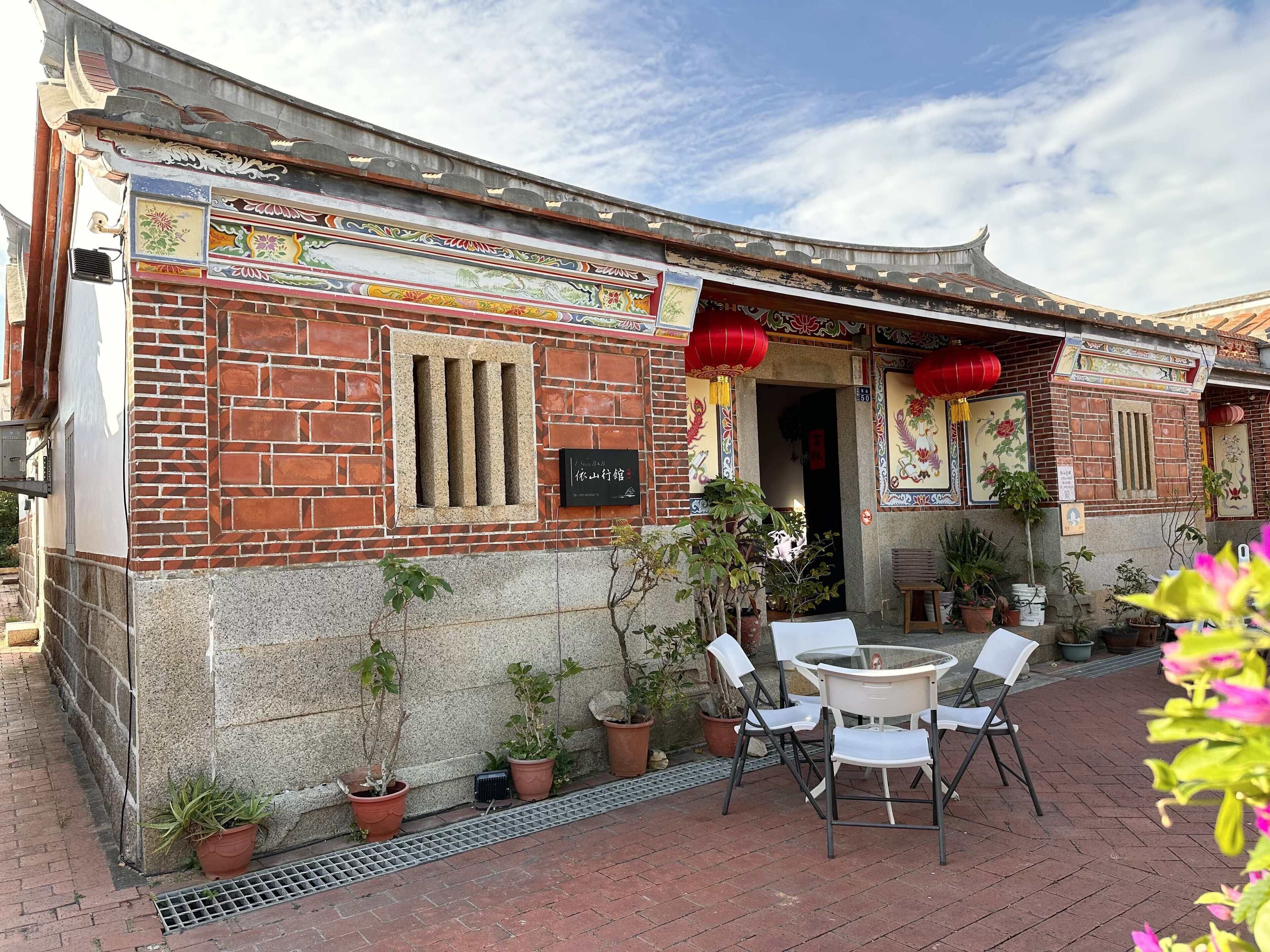 A traditional building with a tiled facade, a brick lower section, and a tiled upper section. There are potted plants and outdoor seating.