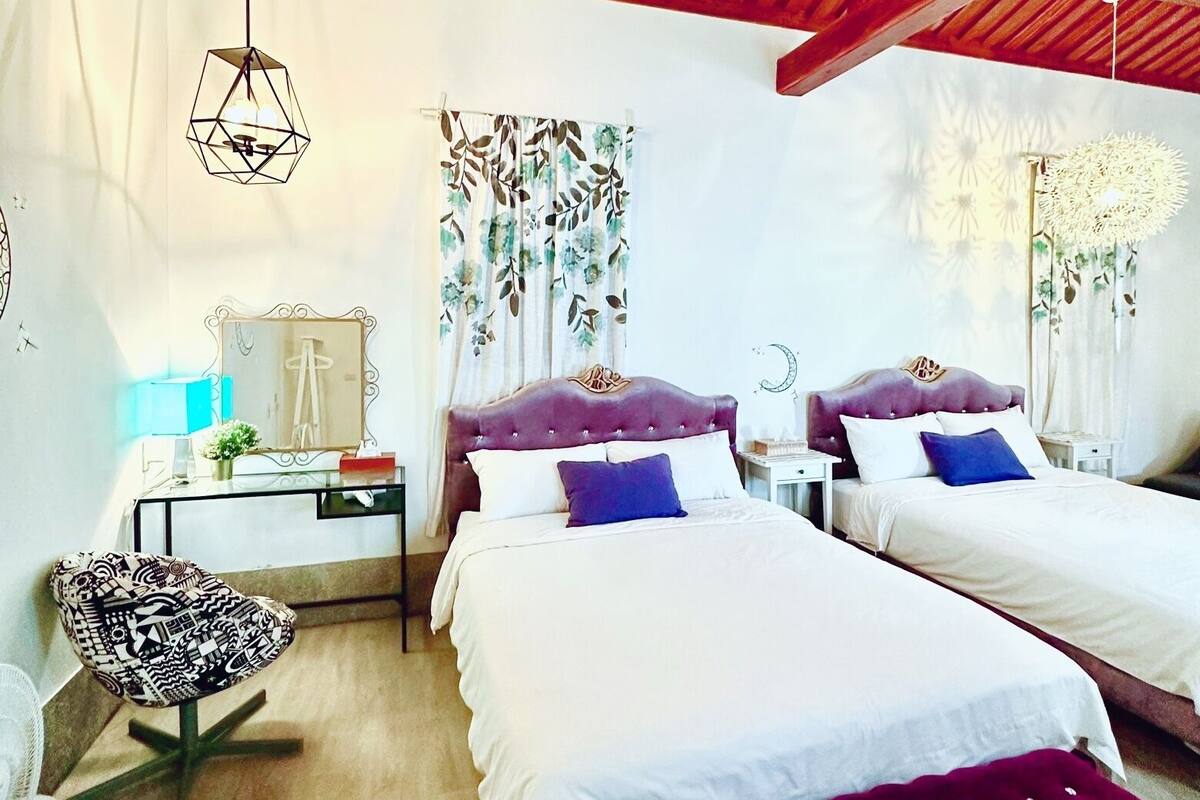 A bedroom with two beds, a wooden ceiling, a geometric light fixture, a floral curtain, and a decorative wall piece.