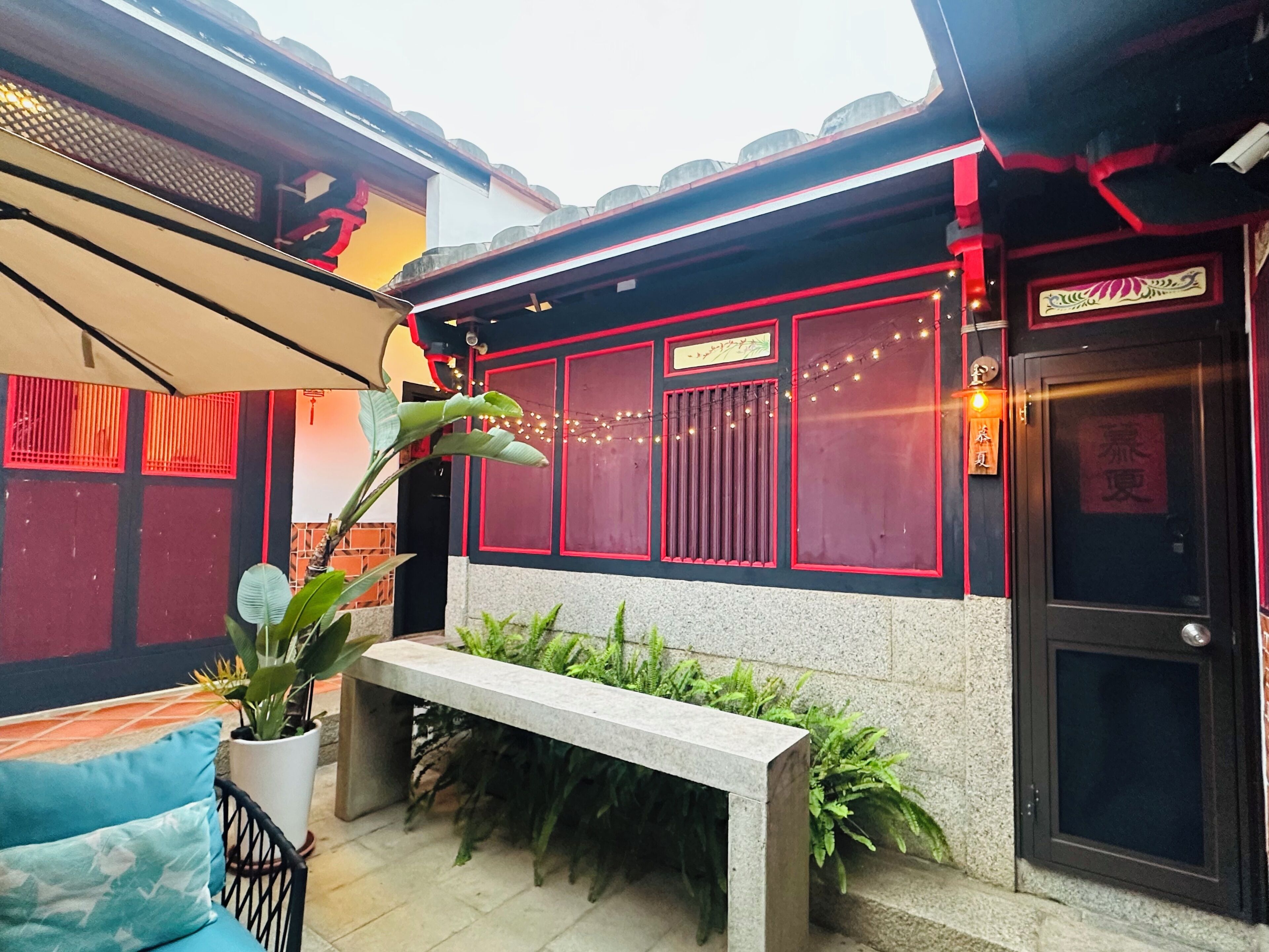 A courtyard with a bench, potted plants, a tiled floor, and a building with red-framed windows and a door.