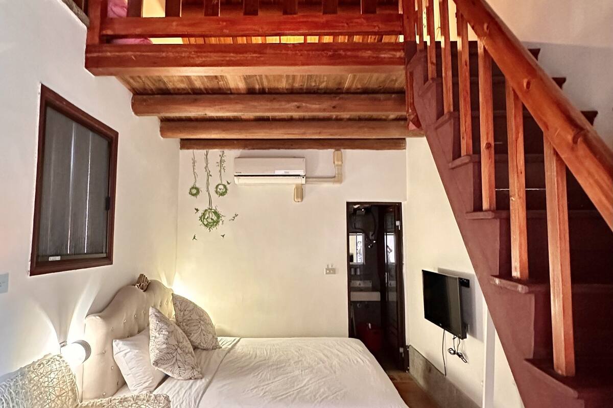 A bedroom with a bed, a wooden staircase, a TV, and a window.