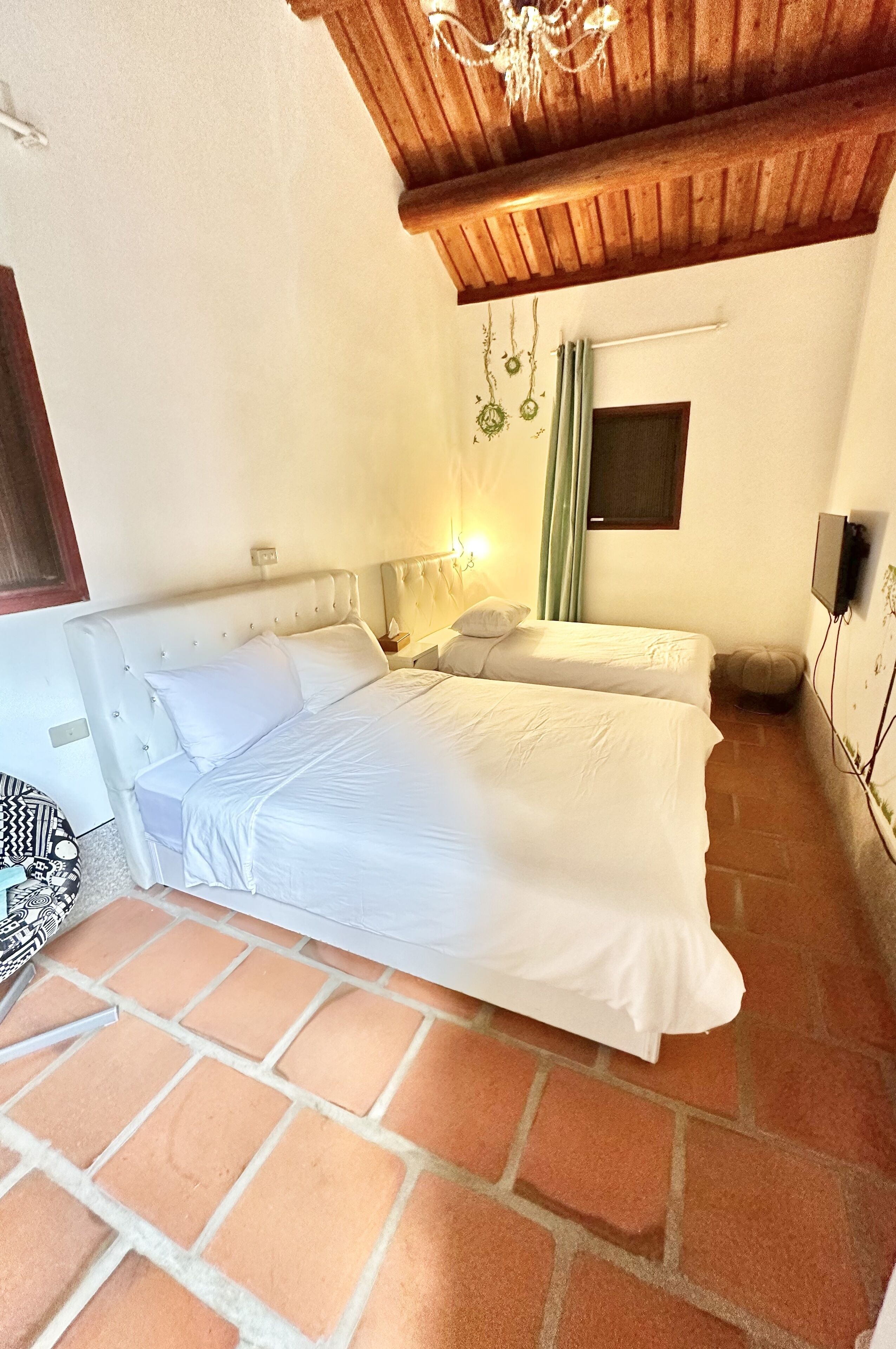 A bedroom with two beds, a wooden ceiling, a small window, and a wall-mounted TV.