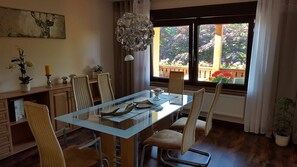 Un coin repas avec une table en verre, des chaises en osier, un lustre et une vue sur un jardin grâce à de grandes fenêtres.