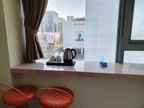 A hotel room with a window view of a cityscape, a kitchen counter with a kettle, cups, and a blue container, and two orange chairs.