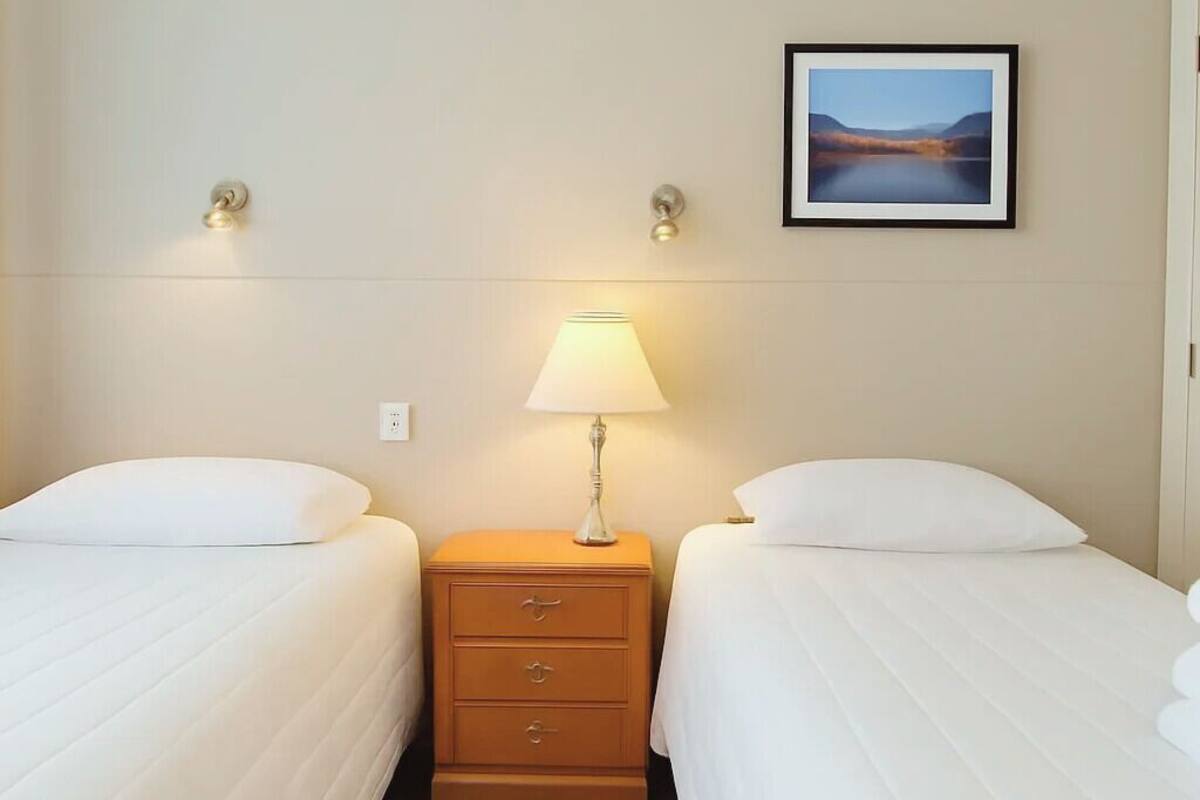 A hotel room with two beds, a nightstand with a lamp, a wall-mounted picture, and a small drawer unit.