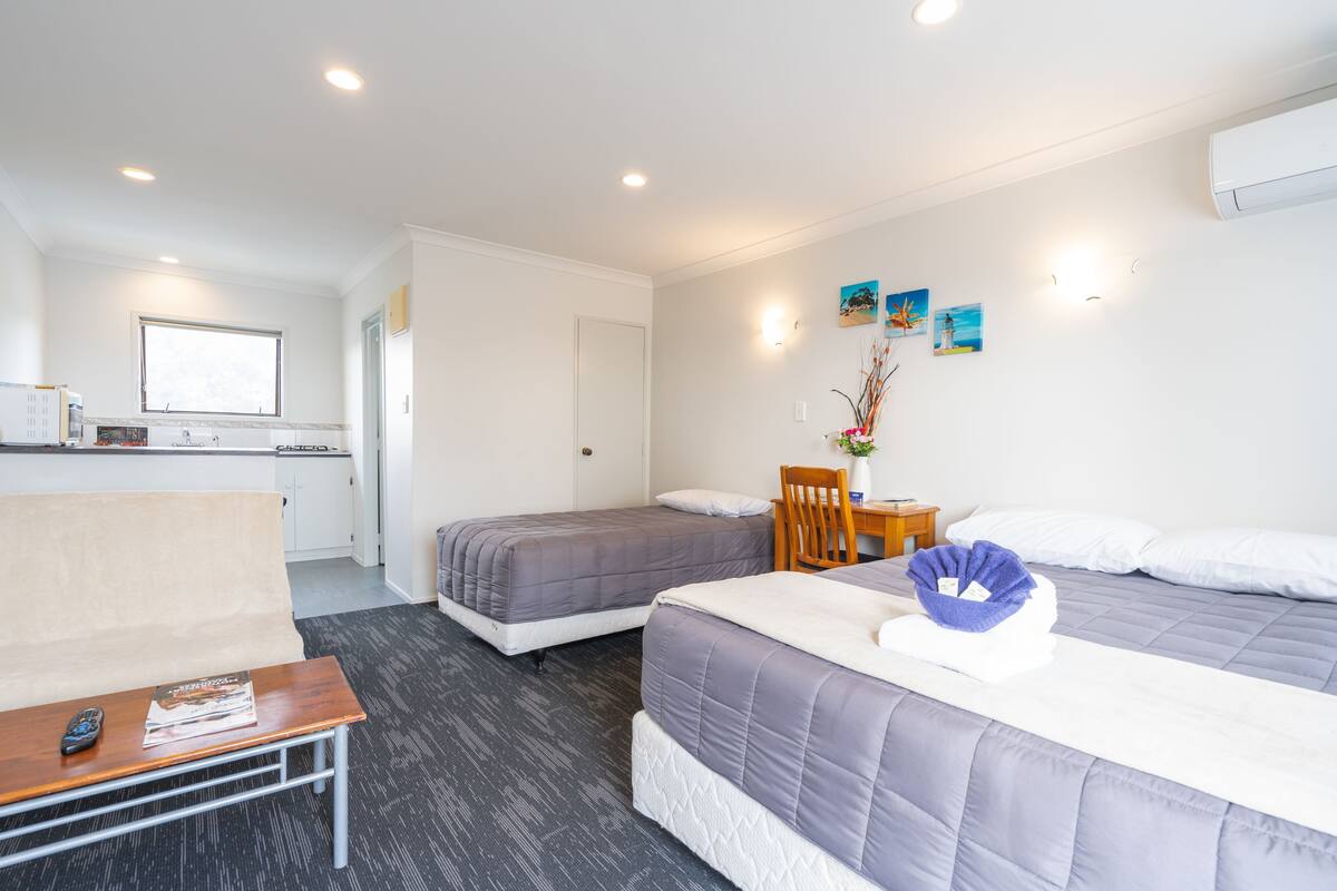 A hotel room with two beds, a small table, a chair, a sofa, a kitchen area, and a window.