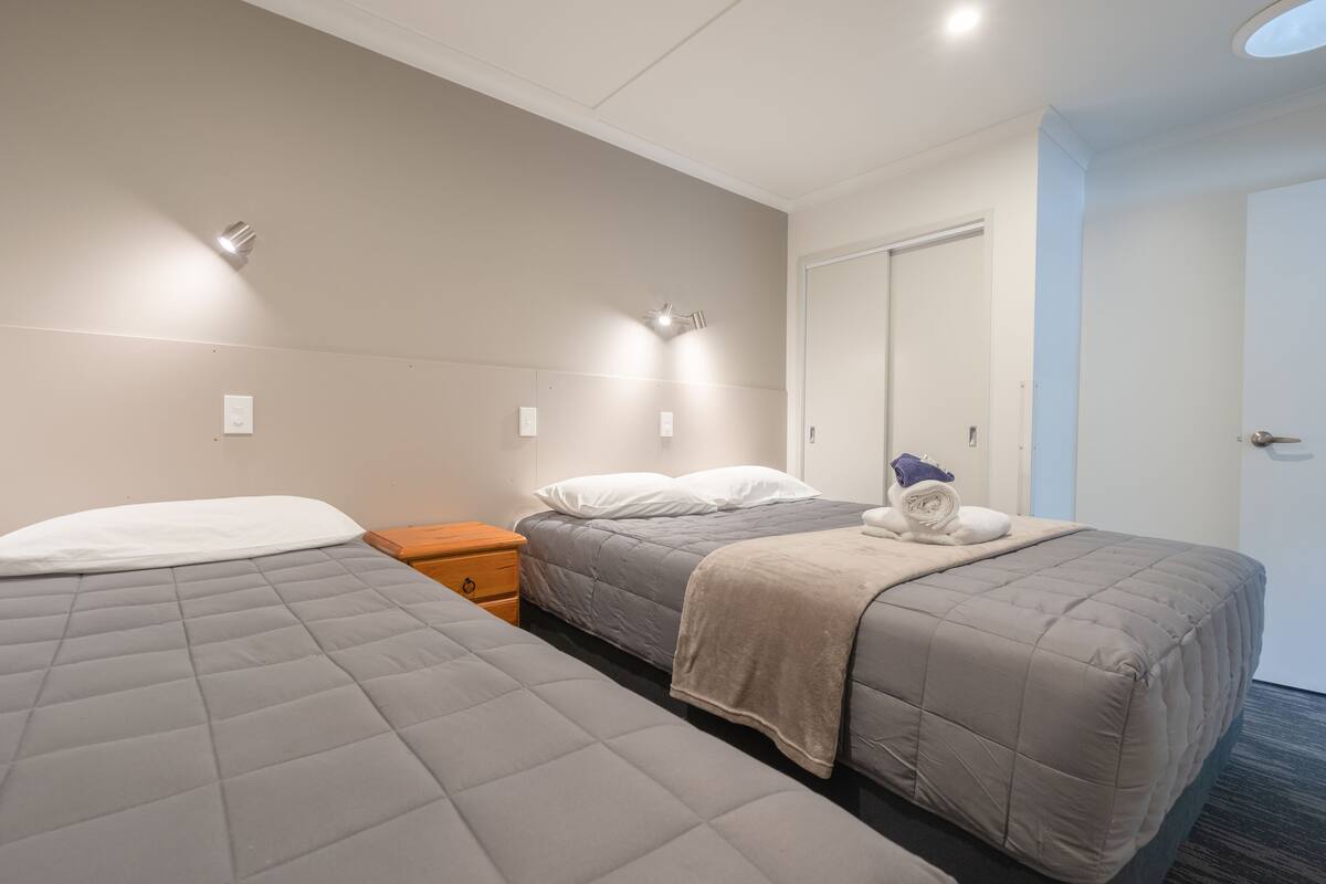 A hotel room with two beds, a wooden nightstand, a white door, and a ceiling light fixture.