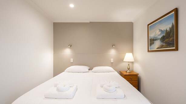 A neatly made bed with two pillows and two towels, a bedside lamp, and a painting of a mountain landscape on the wall. - Pukekohe Motel (Pukekohe)