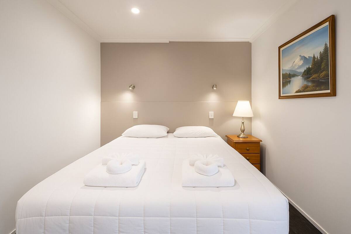 A neatly made bed with two pillows and two towels, a bedside lamp, and a painting of a mountain landscape on the wall.