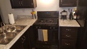 A kitchen with dark cabinets, a stainless steel sink, a stove, and a coffee maker. - Newer Cavco Home in North Phoenix. Near Glendale, Arrowhead and Peoria (Glendale)