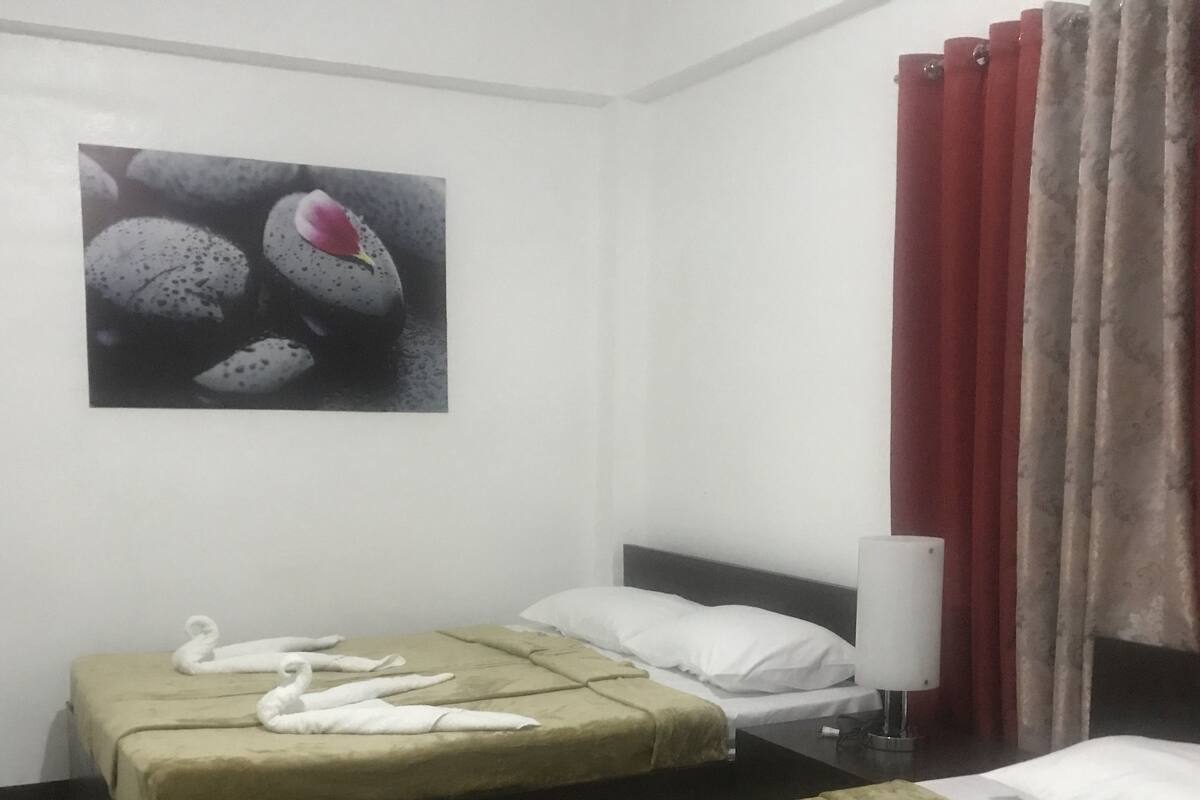 Ein Hotelzimmer mit zwei Betten, jedes mit einer grünen Tagesdecke und weißen Kissen. Darüber hängt ein Gemälde an der Wand, am Fenster ein roter Vorhang und auf dem Nachttisch eine weiße Lampe.
