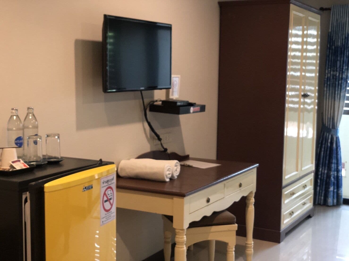 A hotel room with a desk, a television mounted on the wall, a small shelf with a few items, a window with blinds, and a trash bin.