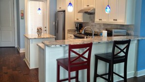 A kitchen with white cabinets, a granite countertop, a stainless steel refrigerator, and two bar stools.