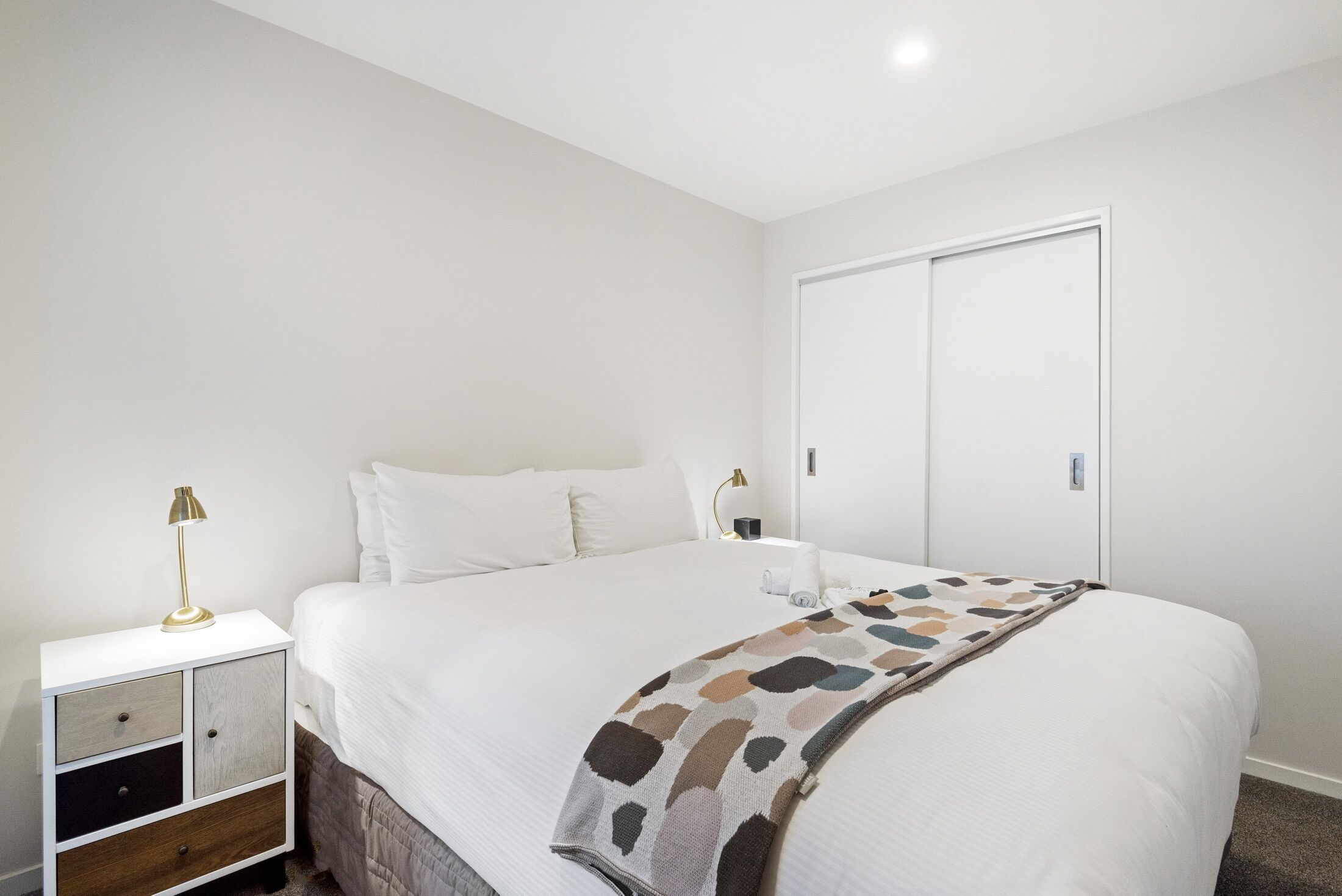 A neatly made bed with a patterned throw, a bedside lamp, and a nightstand with drawers.