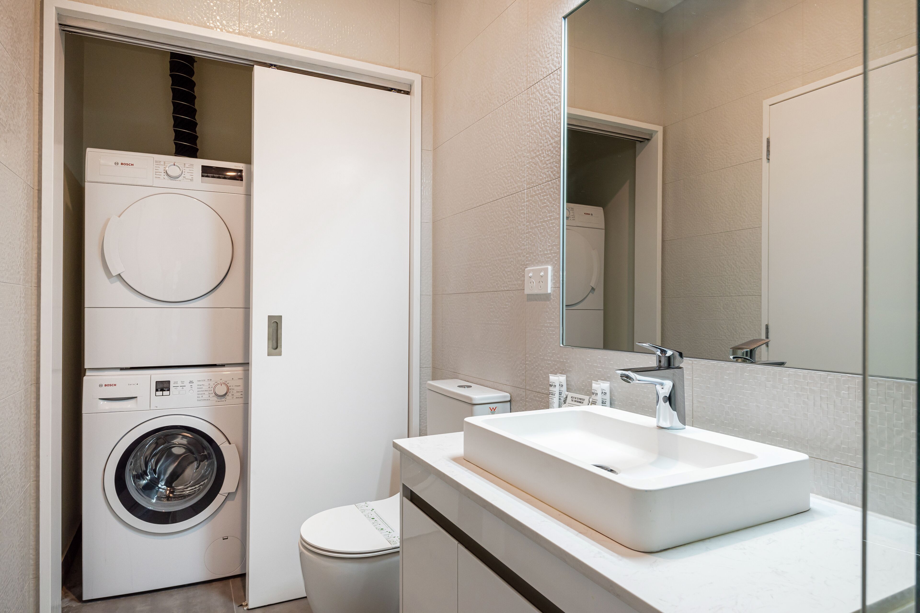 A modern bathroom with a Bosch washing machine, a toilet, a sink with a mirror, and a tiled wall.