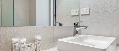 A bathroom with a white sink, two faucets, and a mirror above it. There are three bottles of soap on a wooden tray.