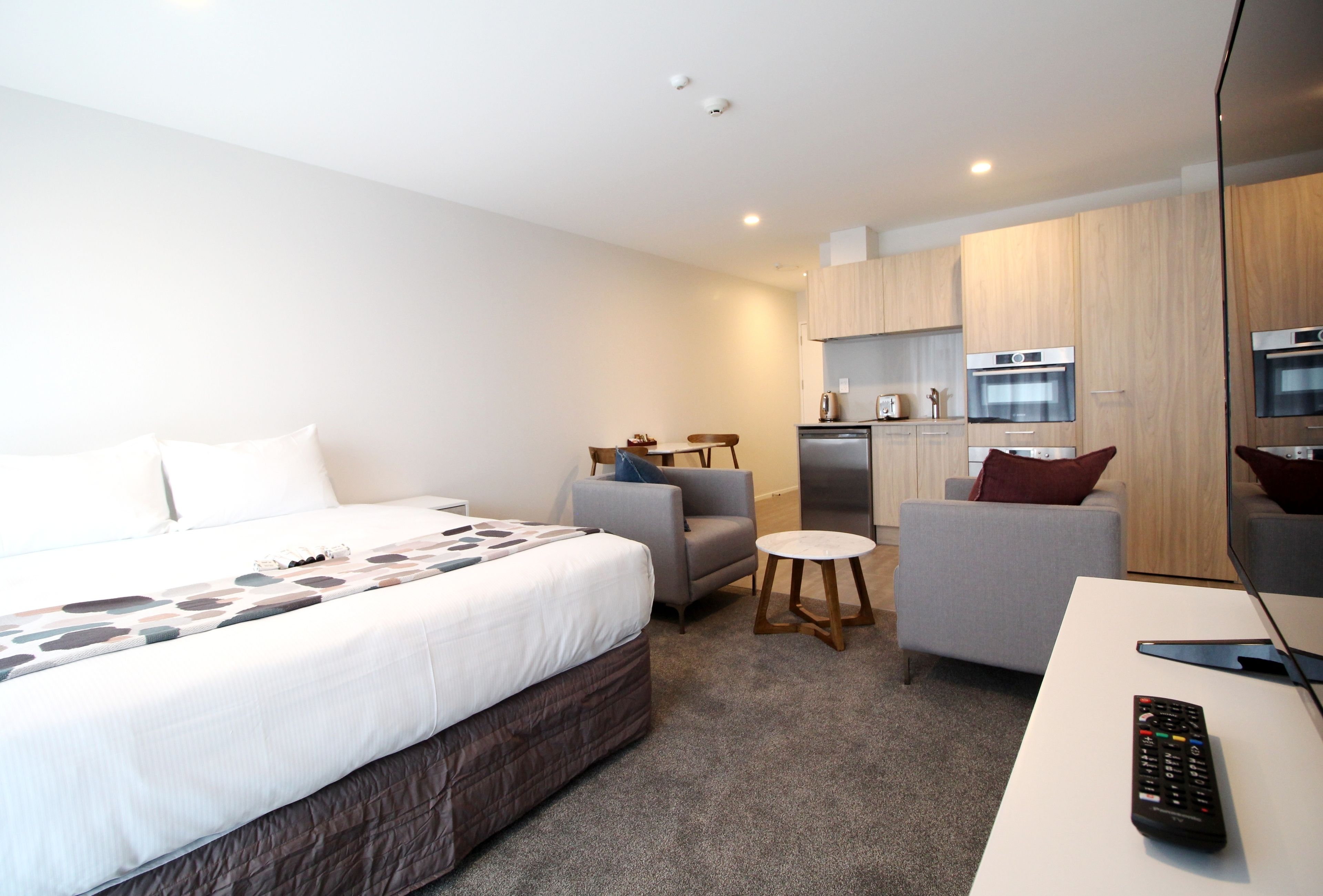 A hotel room with a bed, two armchairs, a small table, a TV, and a kitchenette with a microwave and oven.