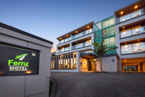 Un edificio moderno con un letrero que indica Fernz Motel y Apartamentos, con varios pisos, ventanas amplias y una entrada bien iluminada.
