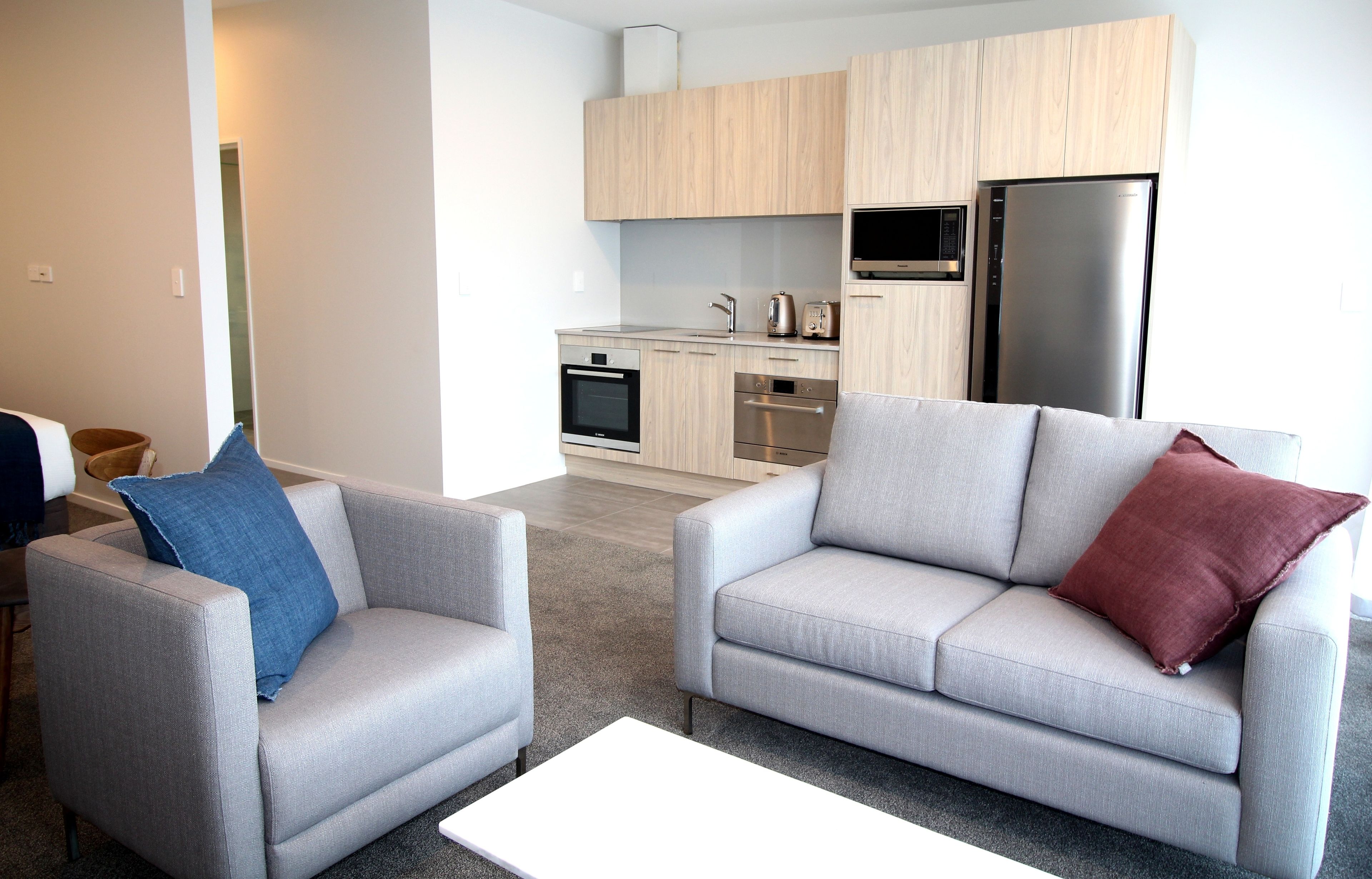 A modern living room with two grey sofas, a white coffee table, and a kitchen area with wooden cabinets, a microwave, and a refrigerator.