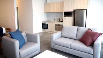 A modern living room with two grey sofas, a white coffee table, and a kitchen area with wooden cabinets, a microwave, and a refrigerator.