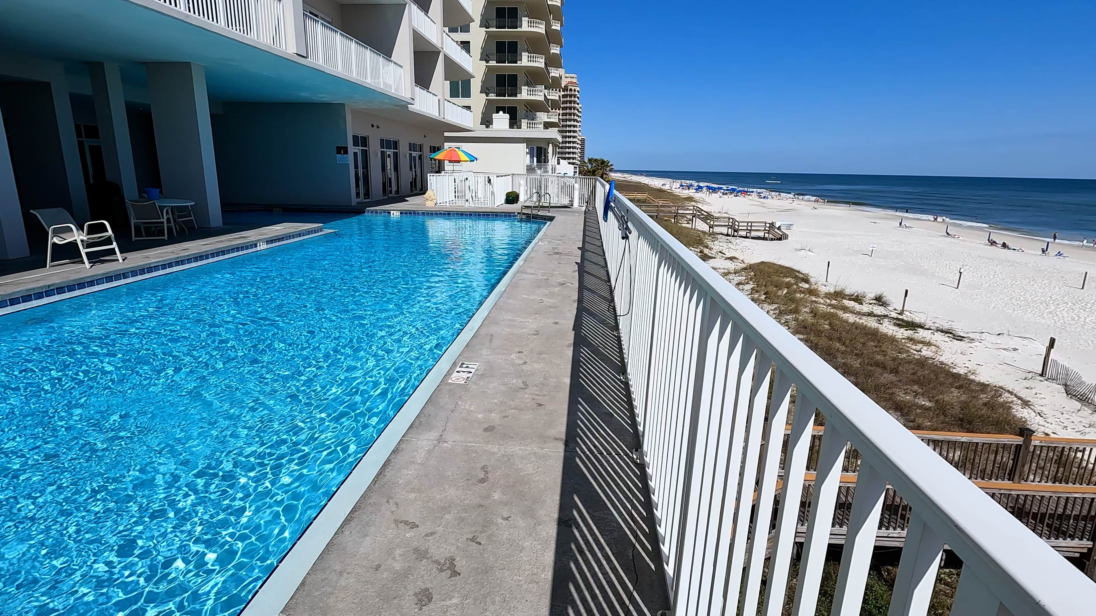 A swimming pool adjacent to a beach, with a multi-story building in the background.