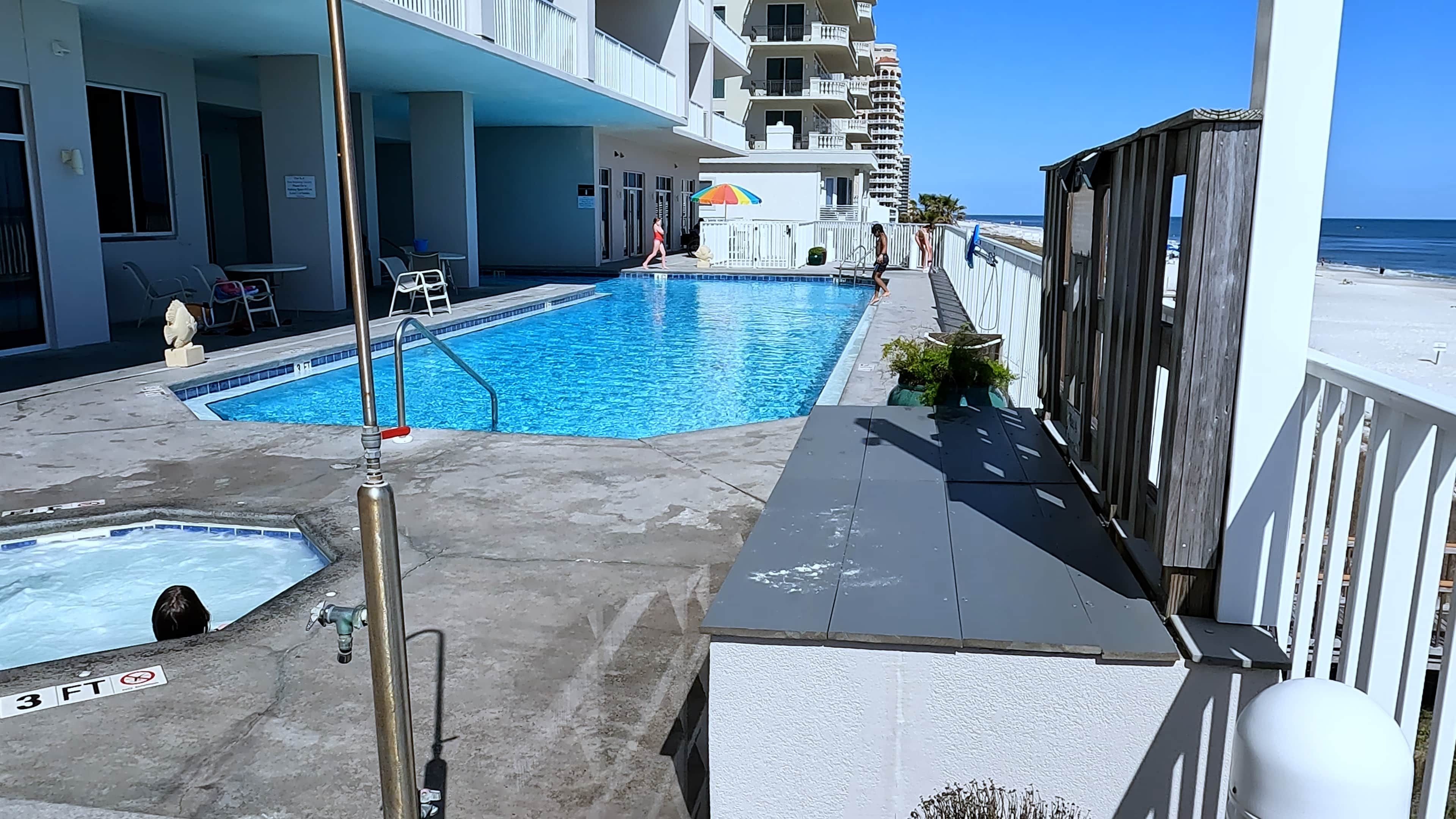 A swimming pool area with a small pool and a larger pool, surrounded by a building with balconies and a view of the beach.