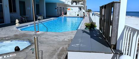 A swimming pool area with a small pool and a larger pool, surrounded by a building with balconies and a view of the beach.