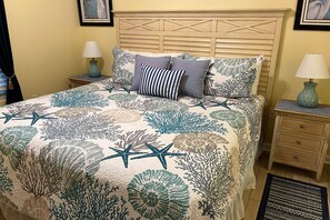 Ein Bett mit Blumen- und Seesternmotiv auf der Tagesdecke, zwei Nachttischlampen, ein hölzernes Kopfteil und ein Nachttisch mit einer Schublade.