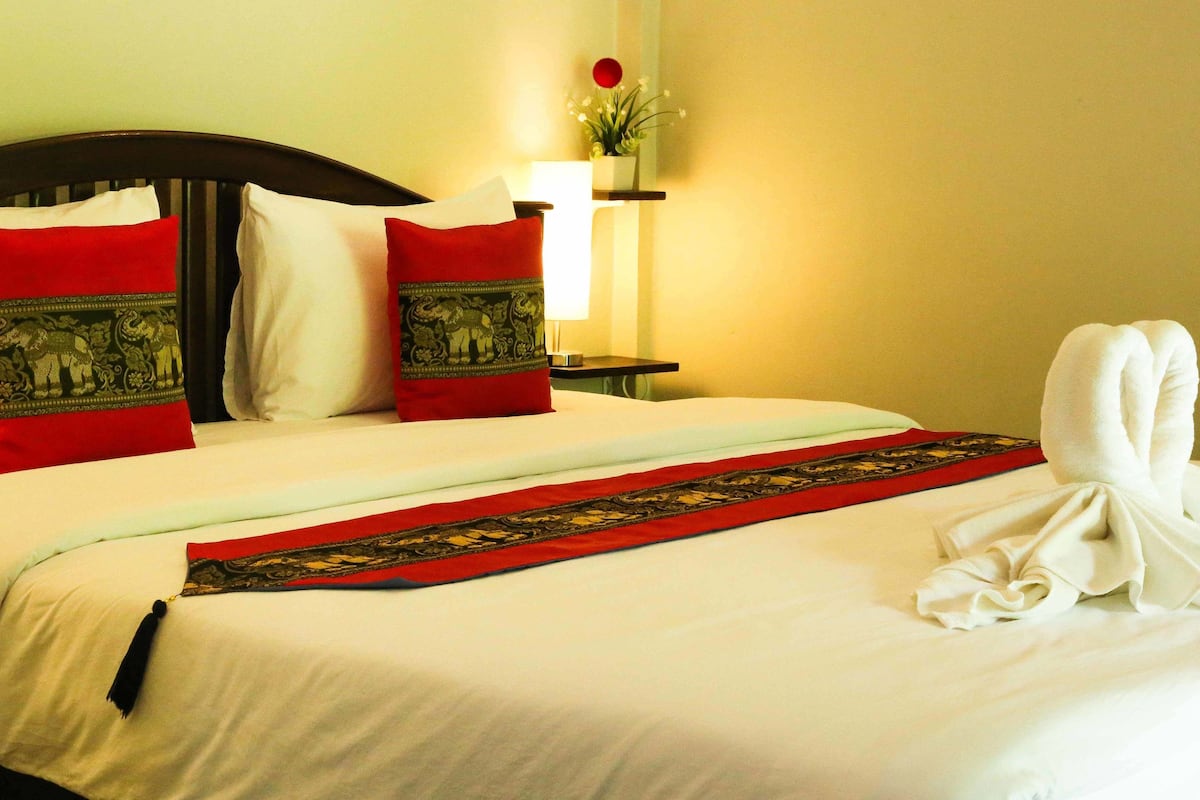A neatly made bed with red and white pillows, a decorative red and black headboard, a white towel on the bed, and a bedside table with a lamp and a small plant.