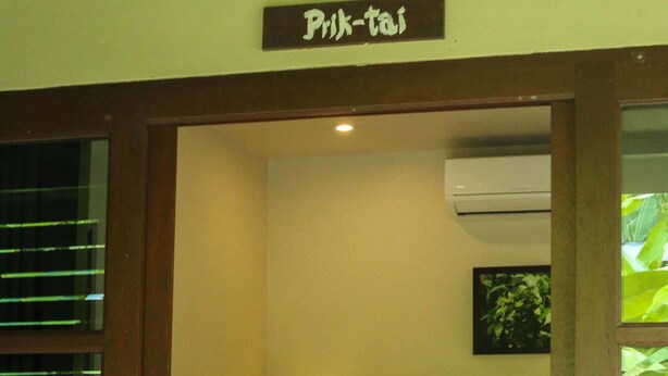 A hotel room with a bed, a nightstand, a lamp, and a sign above the door reading "Prik-tai". - Bang Po Hut (Koh Samui)