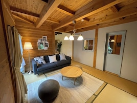A cozy room with a dark sofa, a round wooden table, and a large ottoman. There are framed pictures on the wall and a small window with a view.