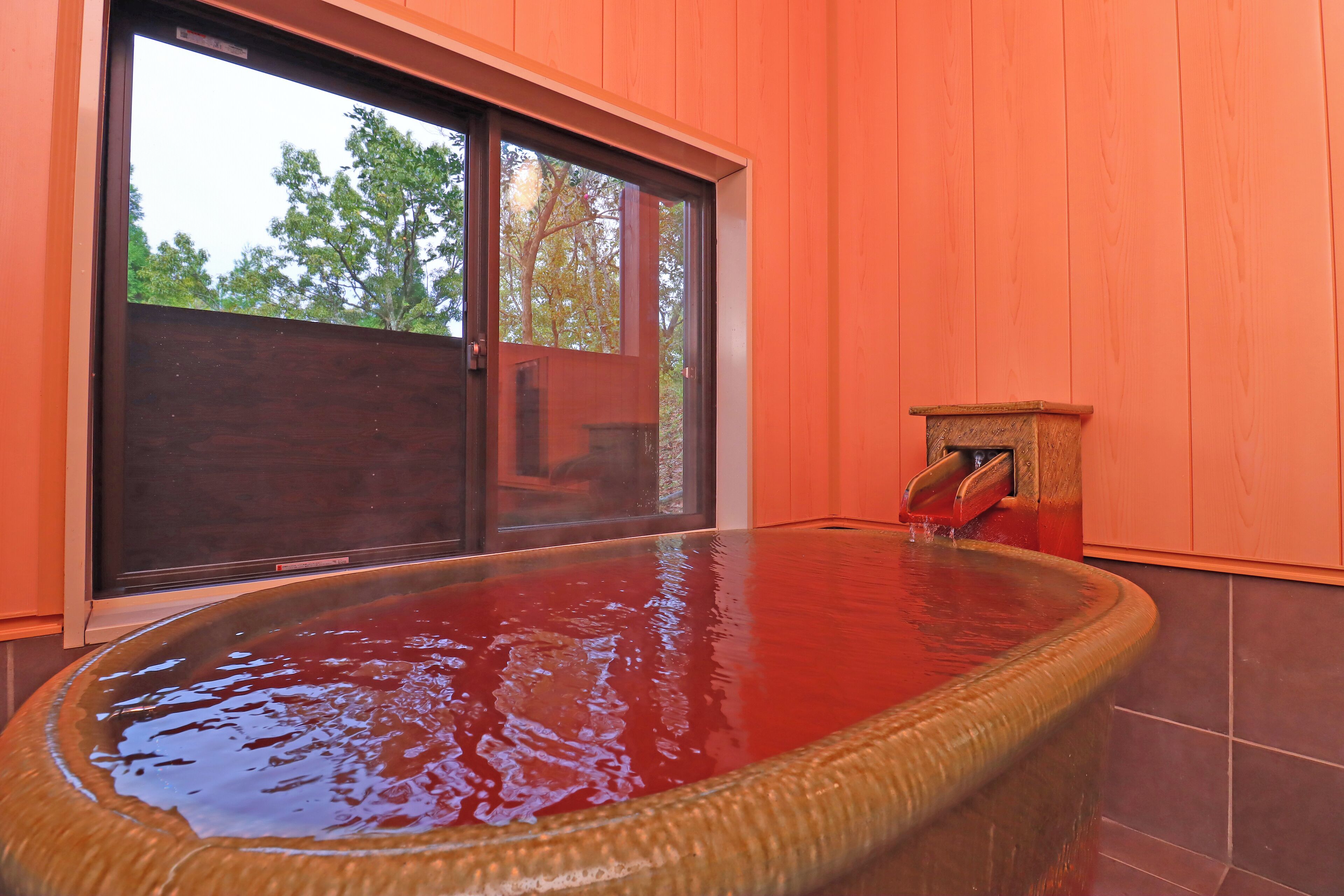 A hot tub with a wooden frame and a small wooden stool beside it, set against a wooden wall with a large window.