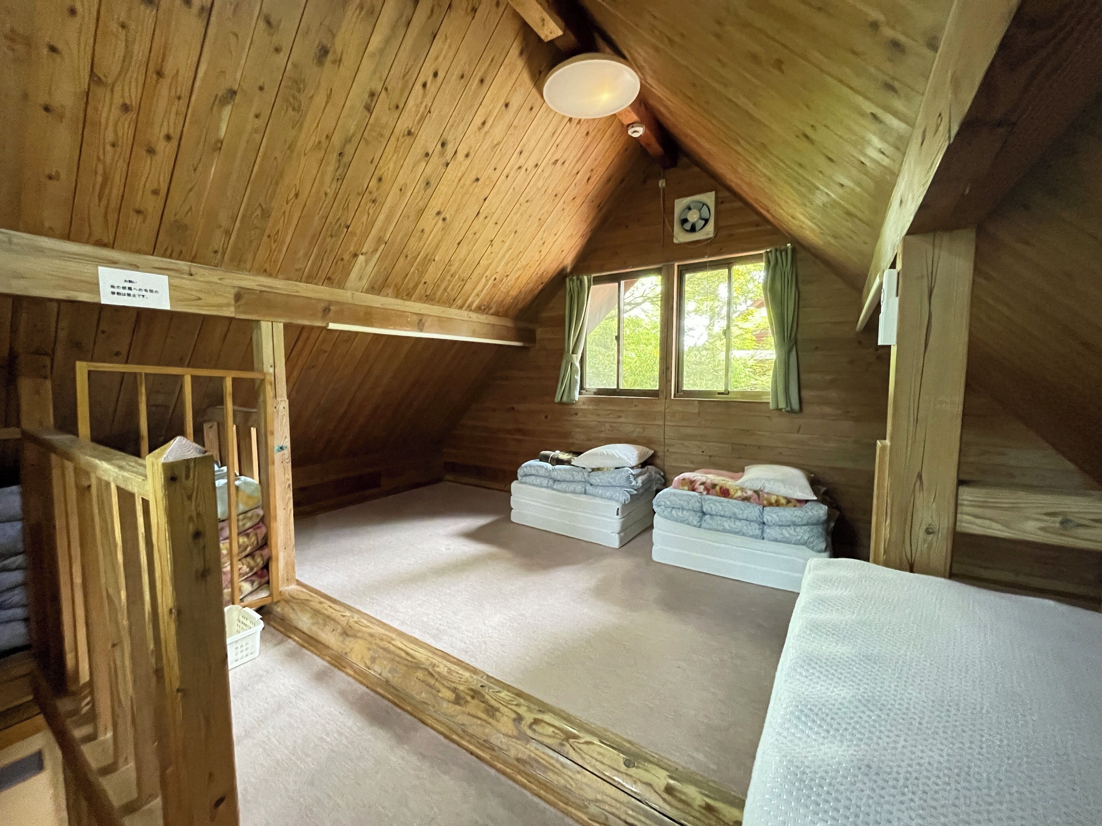 A wooden attic bedroom with two bunk beds, a ceiling fan, and a window with curtains.