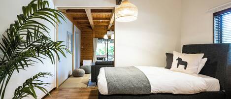 A bedroom with a bed, a large potted plant, wooden flooring, and a hanging light fixture.