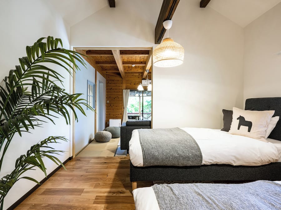 A bedroom with a bed, a large potted plant, wooden flooring, and a hanging light fixture.