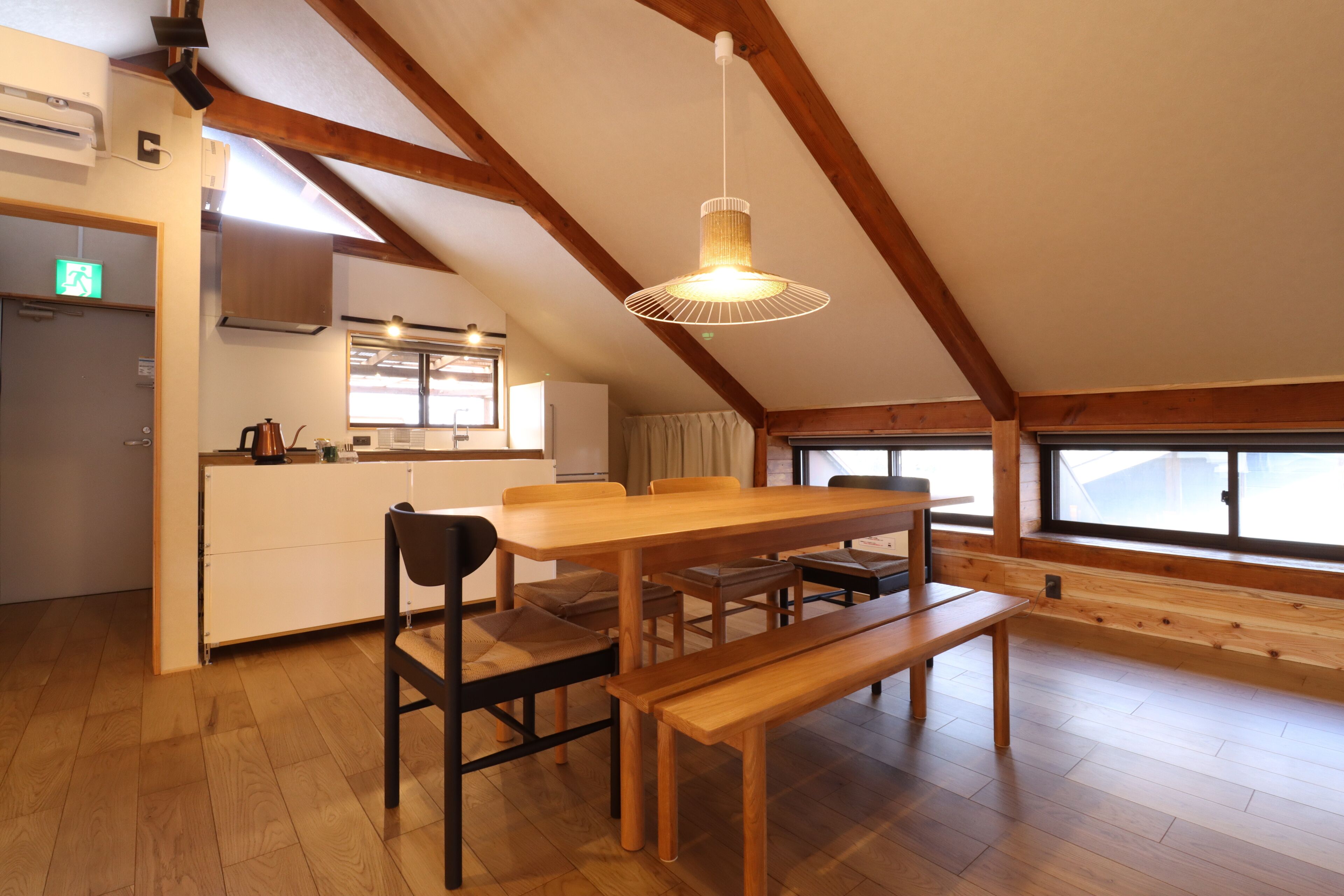 A spacious dining area with a wooden table and benches, a hanging light fixture, and a view through a large window.