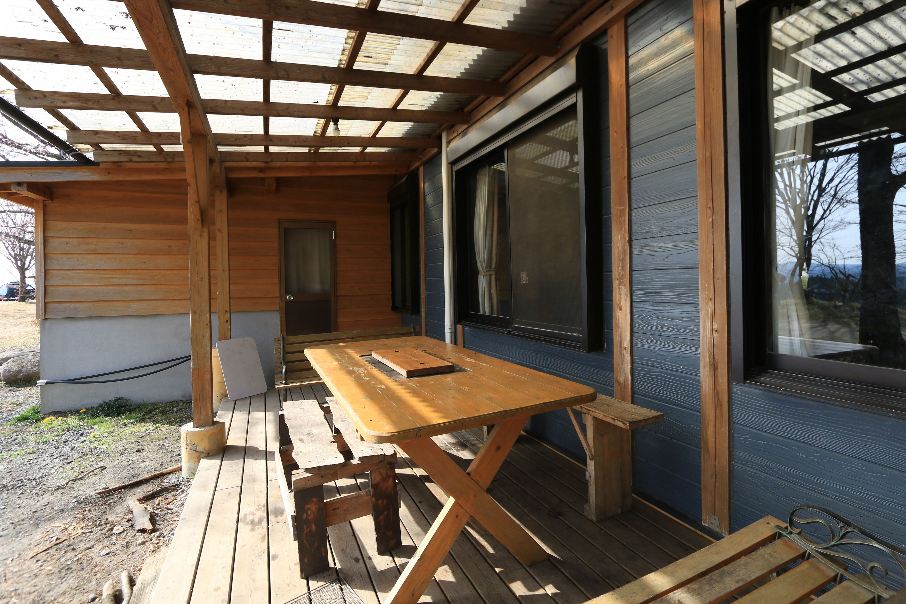 Una terraza de madera con una mesa de picnic y bancos, ventanas grandes y una puerta.
